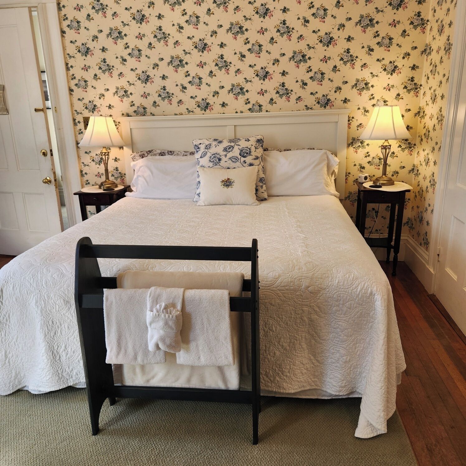 A neatly made bed with white linens, two bedside lamps, and a floral wallpaper pattern.