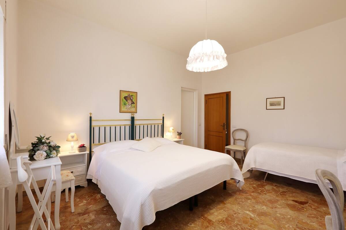 A bedroom with a large bed, a smaller bed, a desk with a chair, a wooden door, and a hanging light fixture.