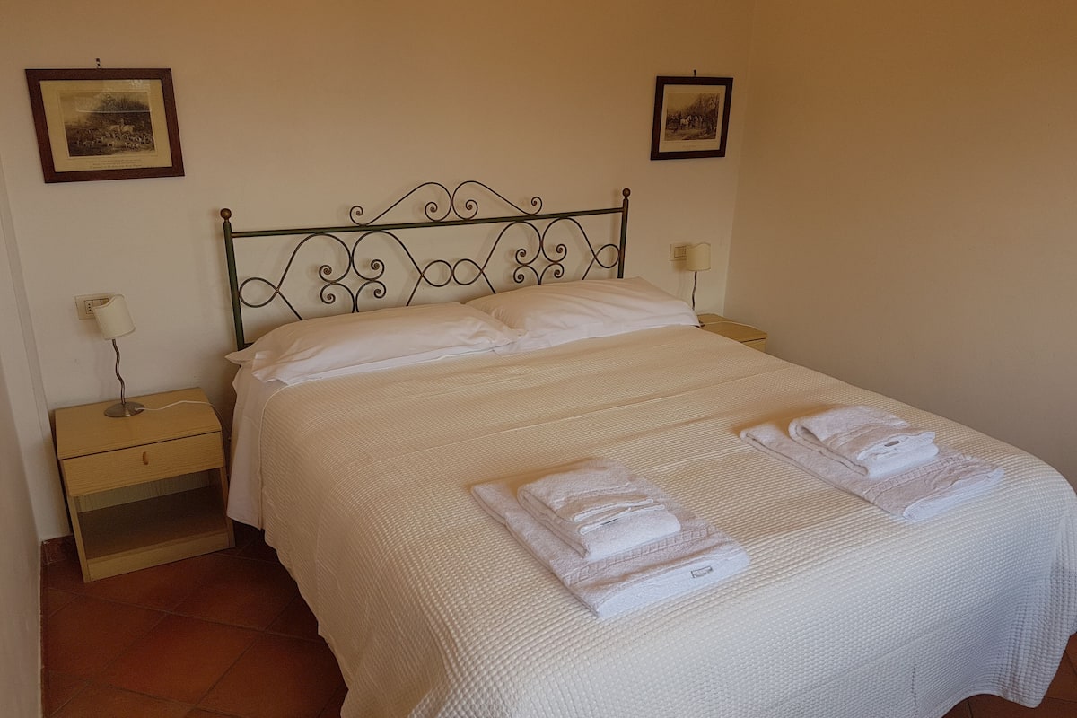 A neatly made bed with white linens and towels, a wooden nightstand with a lamp, and two framed pictures on the wall.