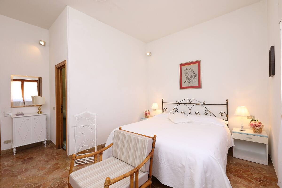 A bedroom with a bed, a chair, a bedside table with a lamp, a dresser, and a mirror.