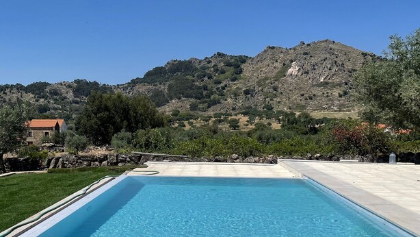 A rectangular swimming pool with clear blue water, surrounded by a stone deck. In the background, there is a mountainous landscape with trees and a building with a red roof.