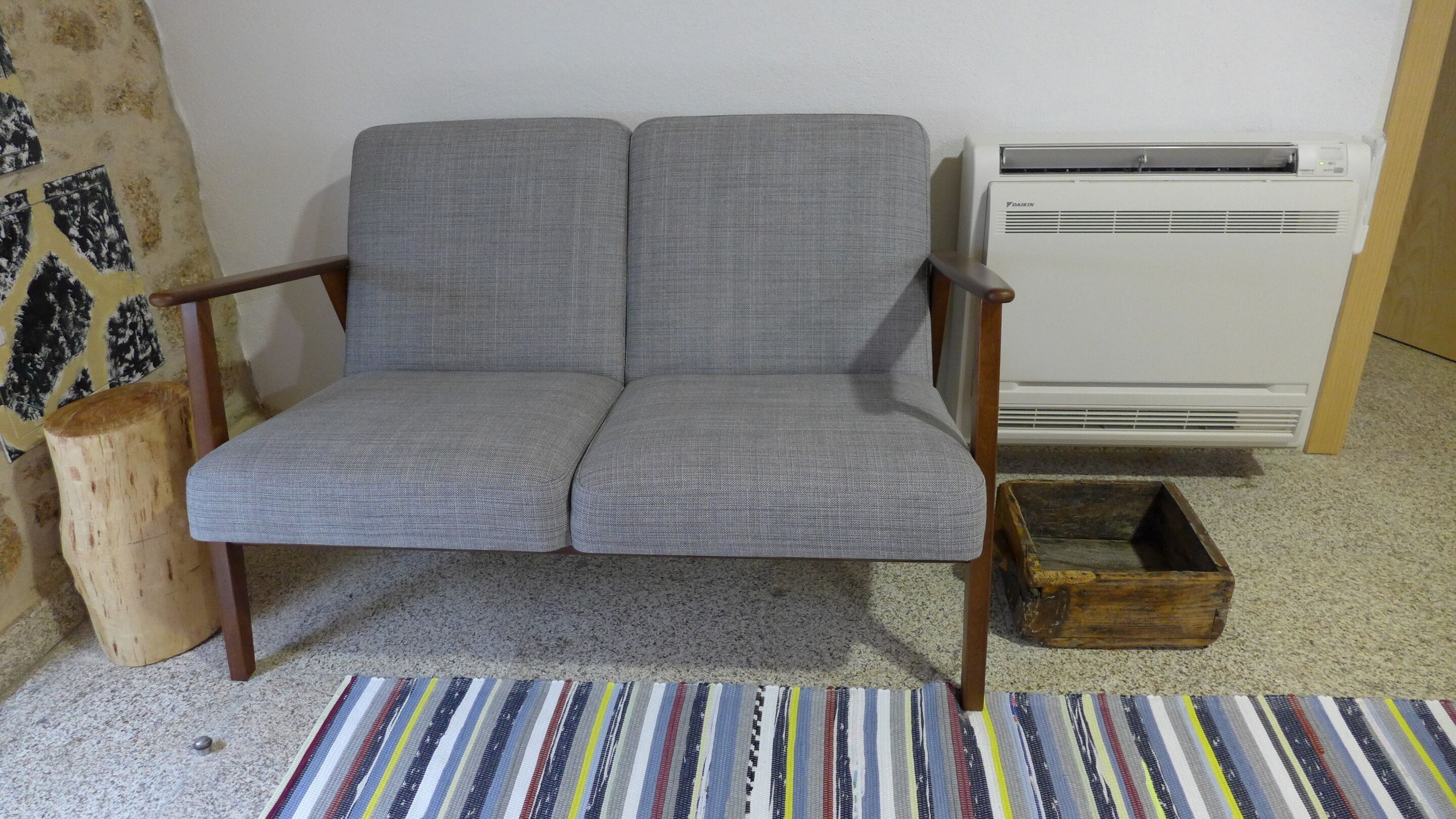 Ein grauer Polsterarmchair mit Holzauflagen, ein kleiner Holzschemel und eine weiße Klimaanlage an einer Wand.