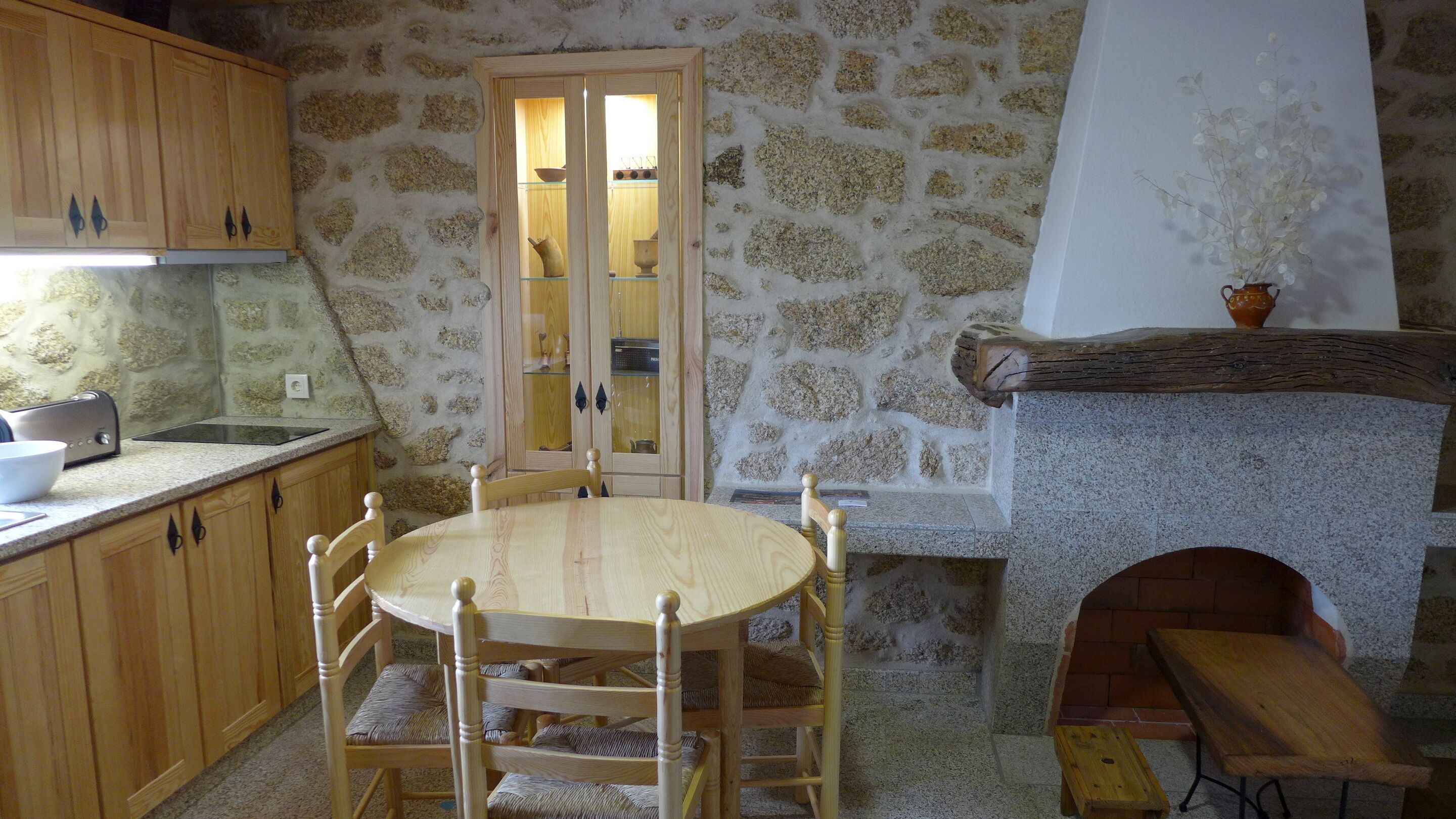 Una cocina rústica con paredes de piedra, armarios de madera, una mesa redonda de madera y un estufa de leña empotrada.