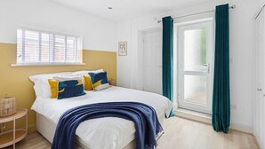 A bedroom with a bed, a window with blinds, a door with curtains, and a small table with a basket. - 84A Albany Road (Cardiff)