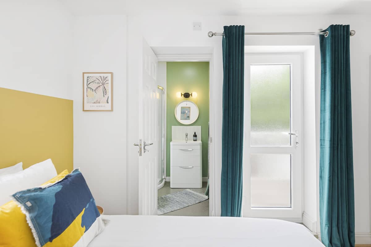 A hotel room with a bed, a bedside table, a nightstand, a wardrobe, a bathroom with a sink and mirror, and a door with curtains.