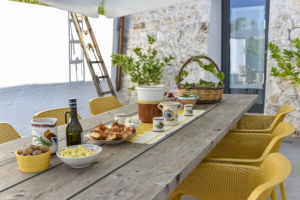 A wooden table set with food and drinks, surrounded by yellow wicker chairs, against a stone wall with a glass door.