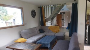 Un salon avec un canapé, un pouf et une table basse. On y trouve un escalier menant à un autre niveau, ainsi qu’une horloge au mur.