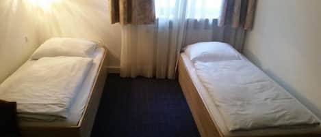 Una habitación pequeña y limpia con dos camas individuales, una ventana con cortinas y una lámpara fijada a la pared.