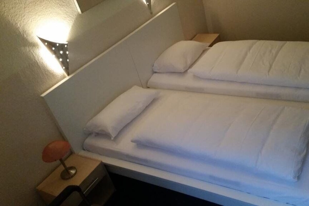 A double bed with white bedding, a bedside table with a lamp, and a wall-mounted light fixture.