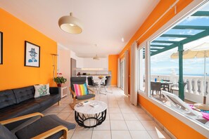 Un salon moderne avec un canapé, des fauteuils et une table basse. On aperçoit une cuisine au fond et un balcon avec des sièges d’extérieur.