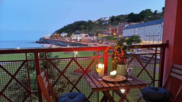 A balcony with a table set for two, overlooking a coastal town with buildings and a sea view.