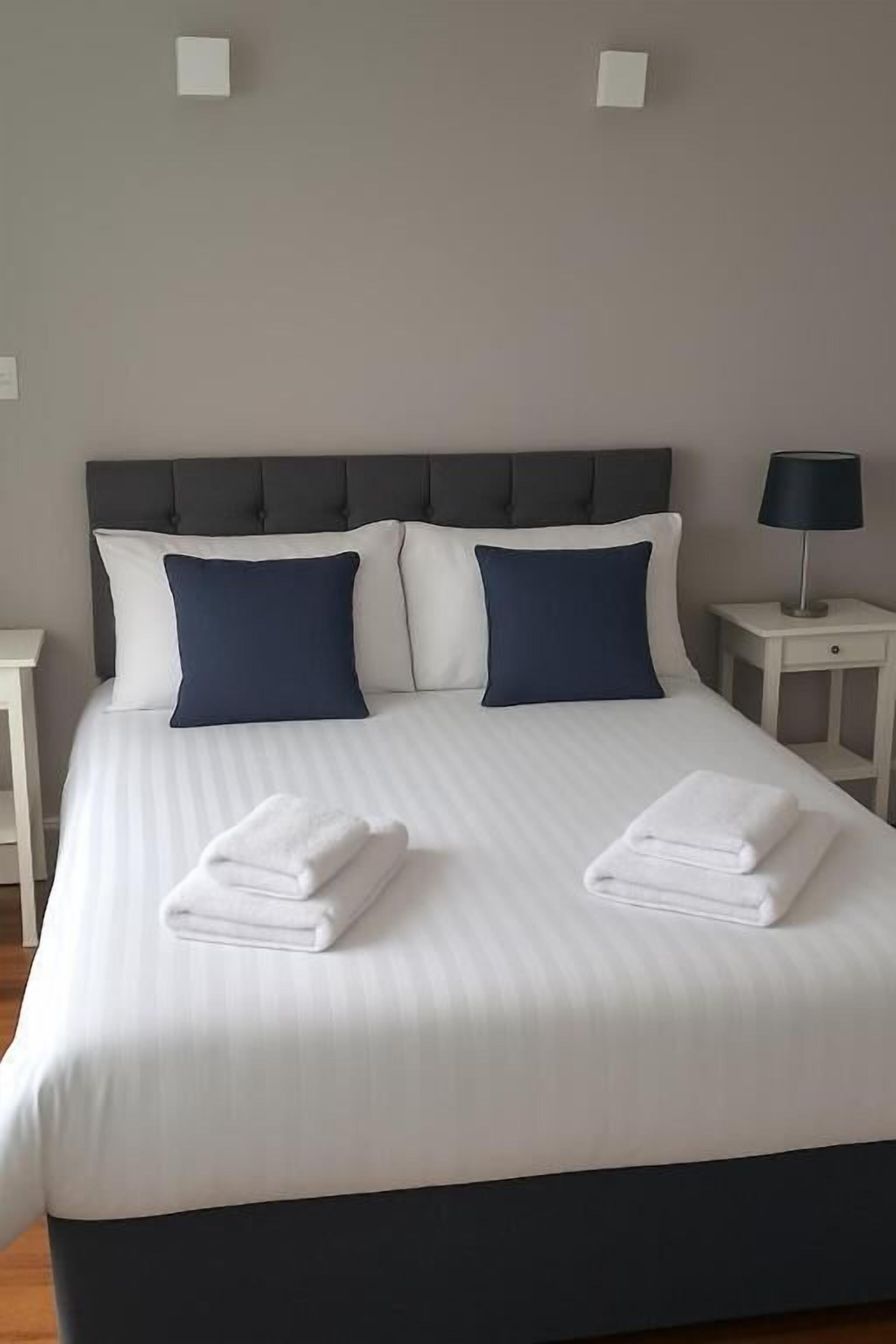 A neatly made bed with white linens and two folded towels, a grey upholstered headboard, and a bedside table with a lamp.