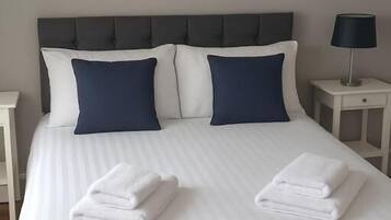 A neatly made bed with white linens and two folded towels, a grey upholstered headboard, and a bedside table with a lamp.