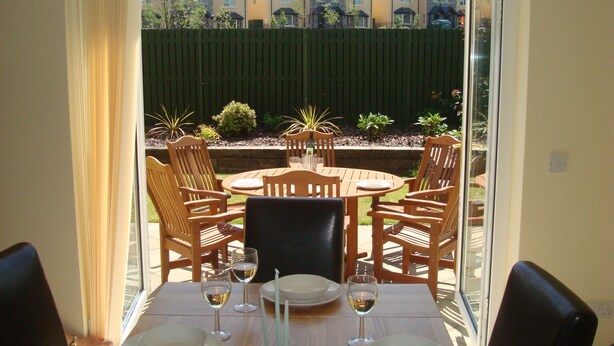 Uma mesa de jantar posta para quatro pessoas, com cadeiras, vista pela janela de uma área residencial e uma cerca verde ao fundo.