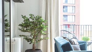 A modern living room with a blue sofa, a potted plant, a wicker basket, and a view of a building through a window. - London Islandic One Excel - O2 - Parking (London)