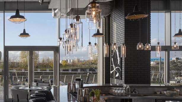 A modern bar area with a long counter, multiple hanging pendant lights, and a view of the city through large windows. - London Islandic One Excel - O2 - Parking (London)