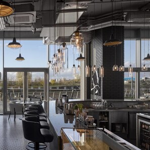 A modern bar area with a long counter, multiple hanging pendant lights, and a view of the city through large windows.