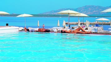 Área de piscina com espreguiçadeiras e guarda-sóis, piscina azul límpida e montanhas ao fundo.
