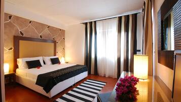 Quarto de hotel com uma cama grande, uma mesa de cabeceira com abajur, um tapete listrado e uma janela com cortinas.