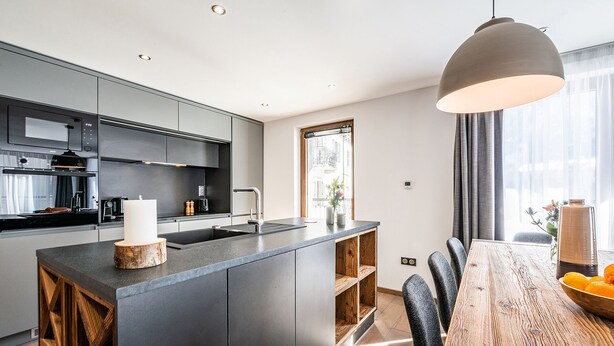 A modern kitchen with dark cabinetry, a central island, and a dining area with a wooden table and chairs. - RAVANEL - Luxury apartment with Mont Blanc views (Chamonix-Mont-Blanc)