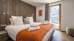 Una camera da letto moderna con una parete in legno decorativa, un letto grande con biancheria da letto bianca, vista sulle montagne attraverso una porta scorrevole e un piccolo angolo salotto.