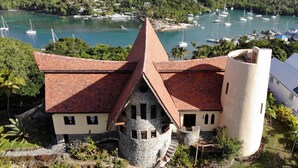 Une maison unique, dotée d’une tour en pierre et d’un toit conique, entourée d’une végétation luxuriante et bordée d’un port de plaisance où sont amarrés des bateaux.