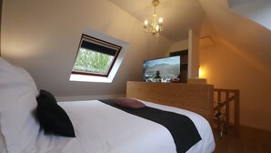 Un dormitorio con cama, televisor en un tocador de madera, candelabro y ventana con vista a árboles.