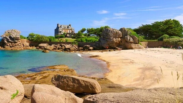 Un paisaje costero con una casa grande sobre un promontorio rocoso, agua azul cristalina y una playa de arena.
