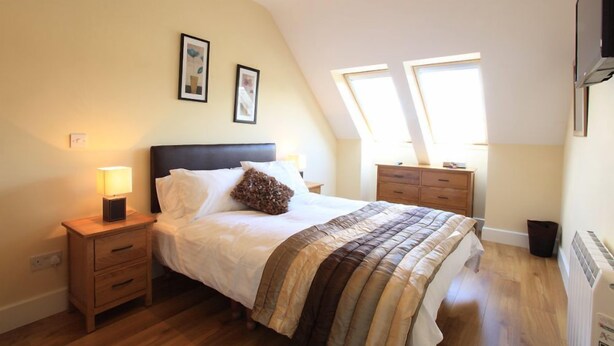 Een slaapkamer met een bed, nachtkastjes, een dressoir en een radiator. Twee ramen zorgen voor natuurlijk licht.