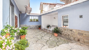 Un patio con suelo de baldosas, plantas en macetas, una zona de estar con sillas y una mesa, y un ventanal con persianas.