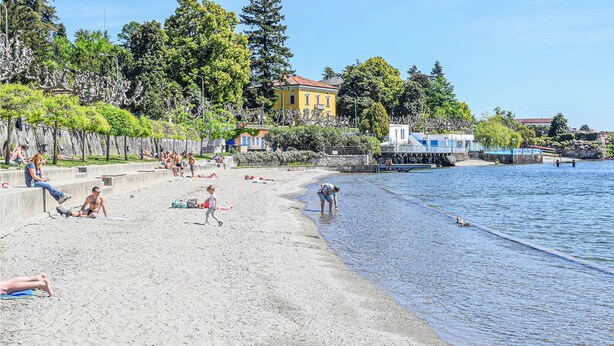 Una playa con gente tomando el sol y nadando, un muelle que se adentra en el agua y edificios al fondo.