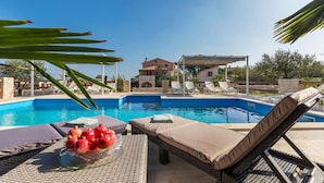 Un espace aménagé au bord de la piscine, avec des chaises longues, une table sur laquelle est posé un panier de fruits, et une vue sur une maison en arrière-plan.