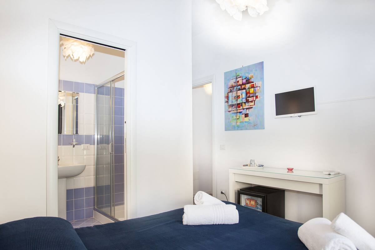 A hotel room with a bed, a desk, a TV, a painting, and a bathroom with a shower and sink.