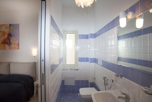 A bathroom with a toilet, sink, and shower. There is a bed and a painting on the wall. - Casa Don Micio (Taormina)