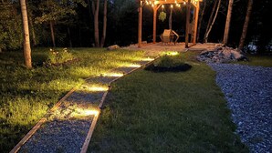Een houten pergola met lichtjes, omgeven door een grindpad en gazon, gelegen in een bebost gebied.