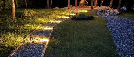 A wooden pergola with string lights, surrounded by a gravel path and grass, set in a wooded area.