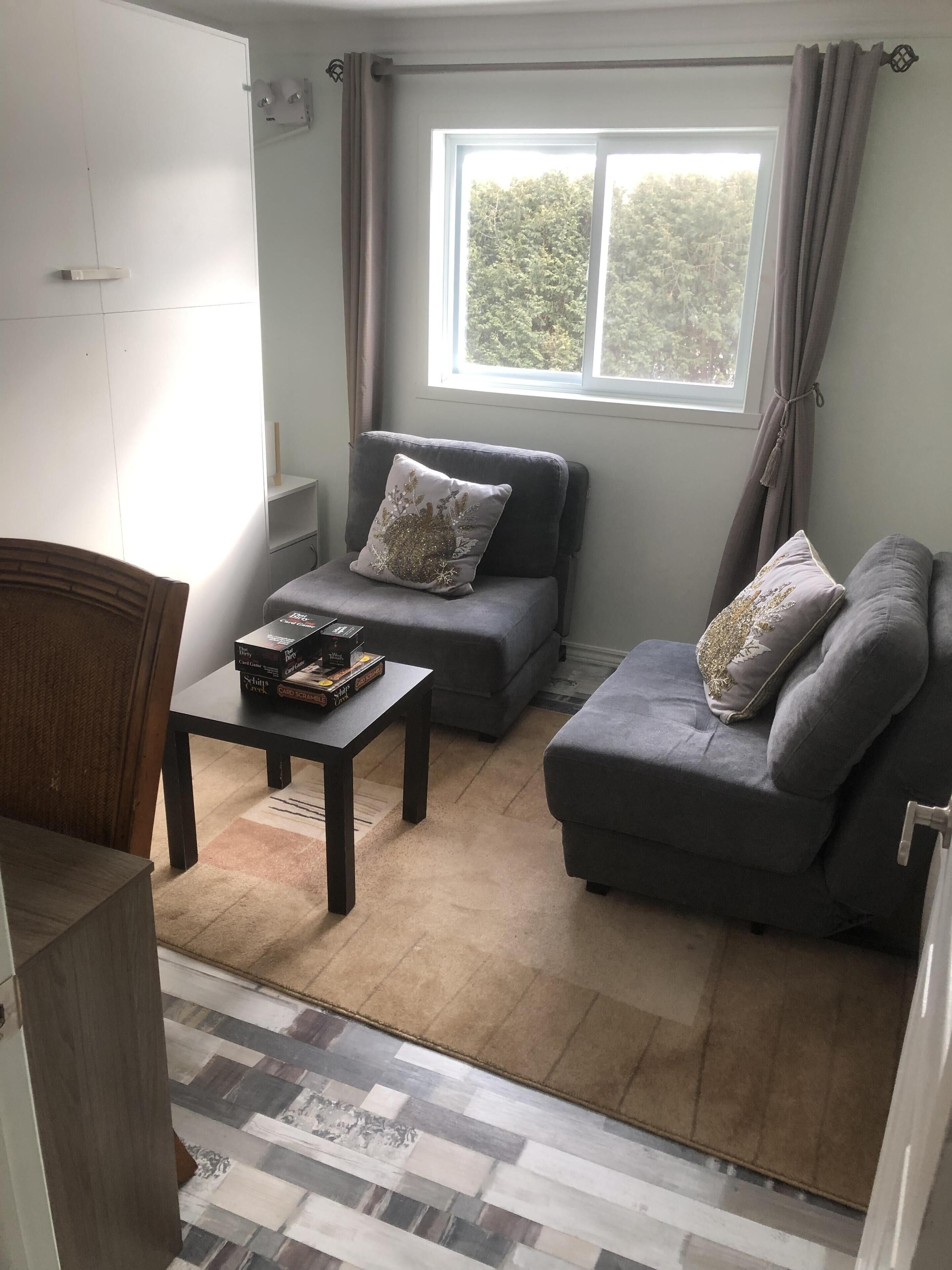 A living room with a sofa set, a coffee table with board games, a window with curtains, and a patterned rug.