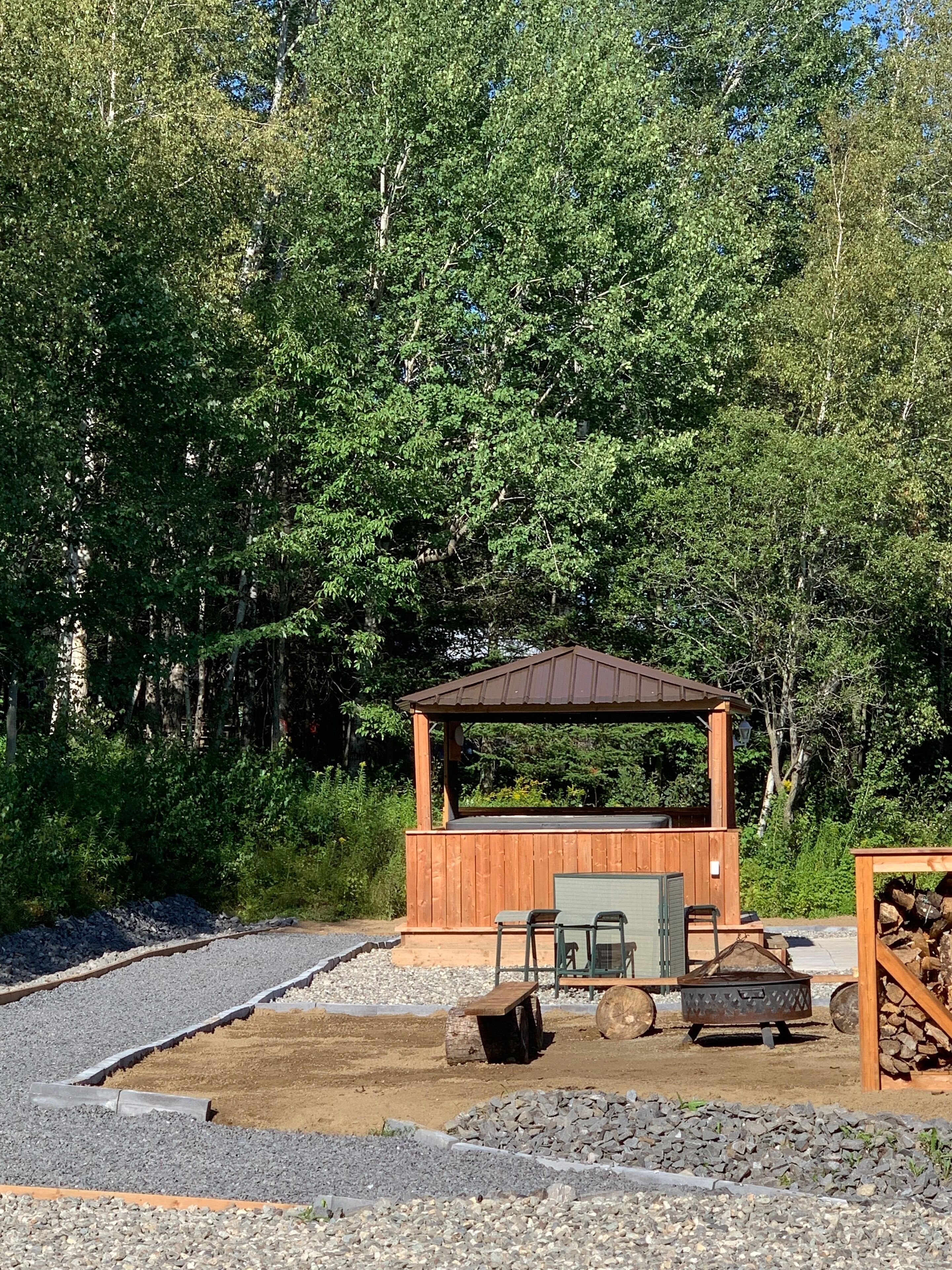 A wooden gazebo with a metal roof, surrounded by gravel and trees.