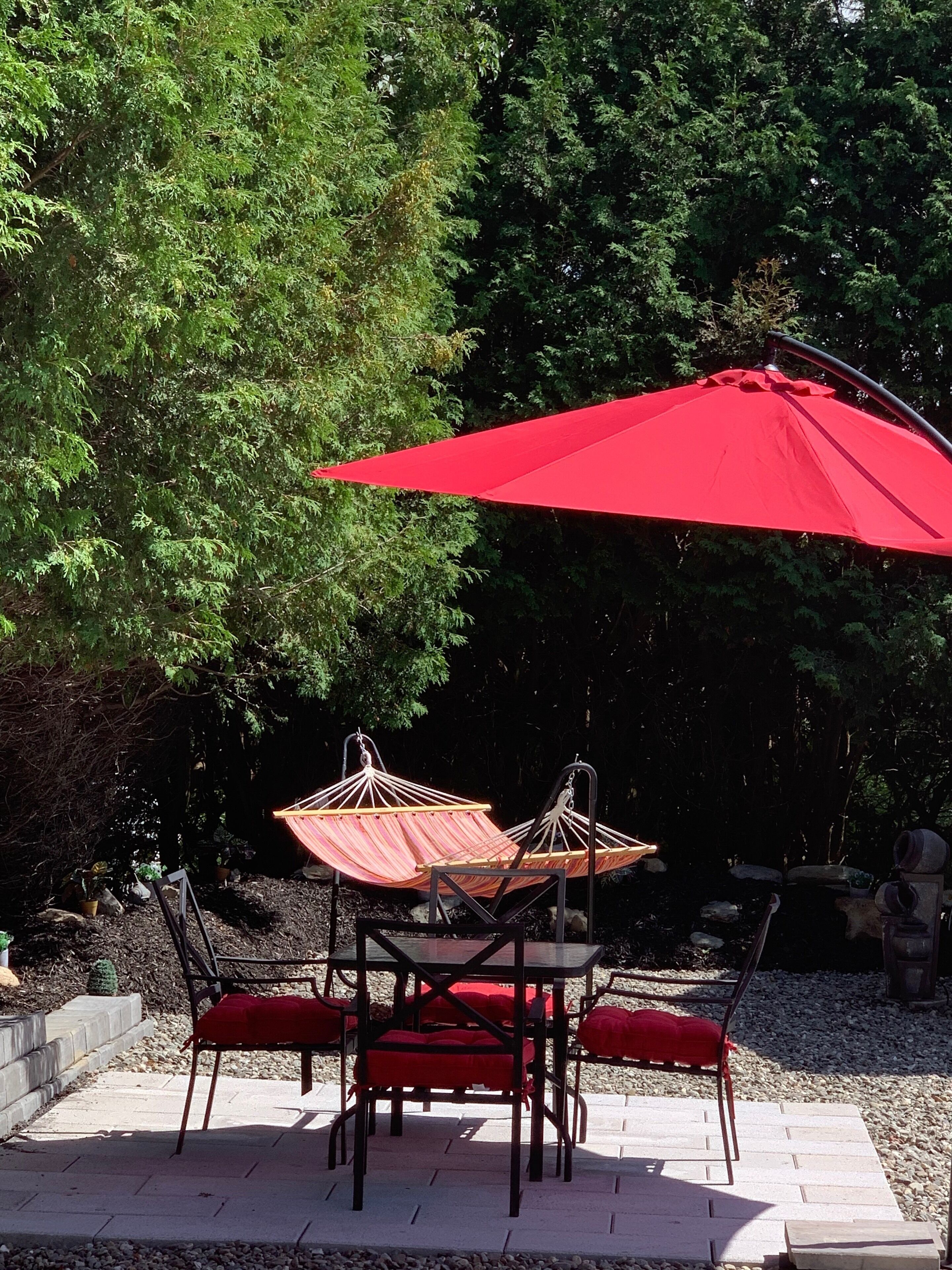 Een terras buiten met een tafel, stoelen, een rode parasol en een hangstoel.