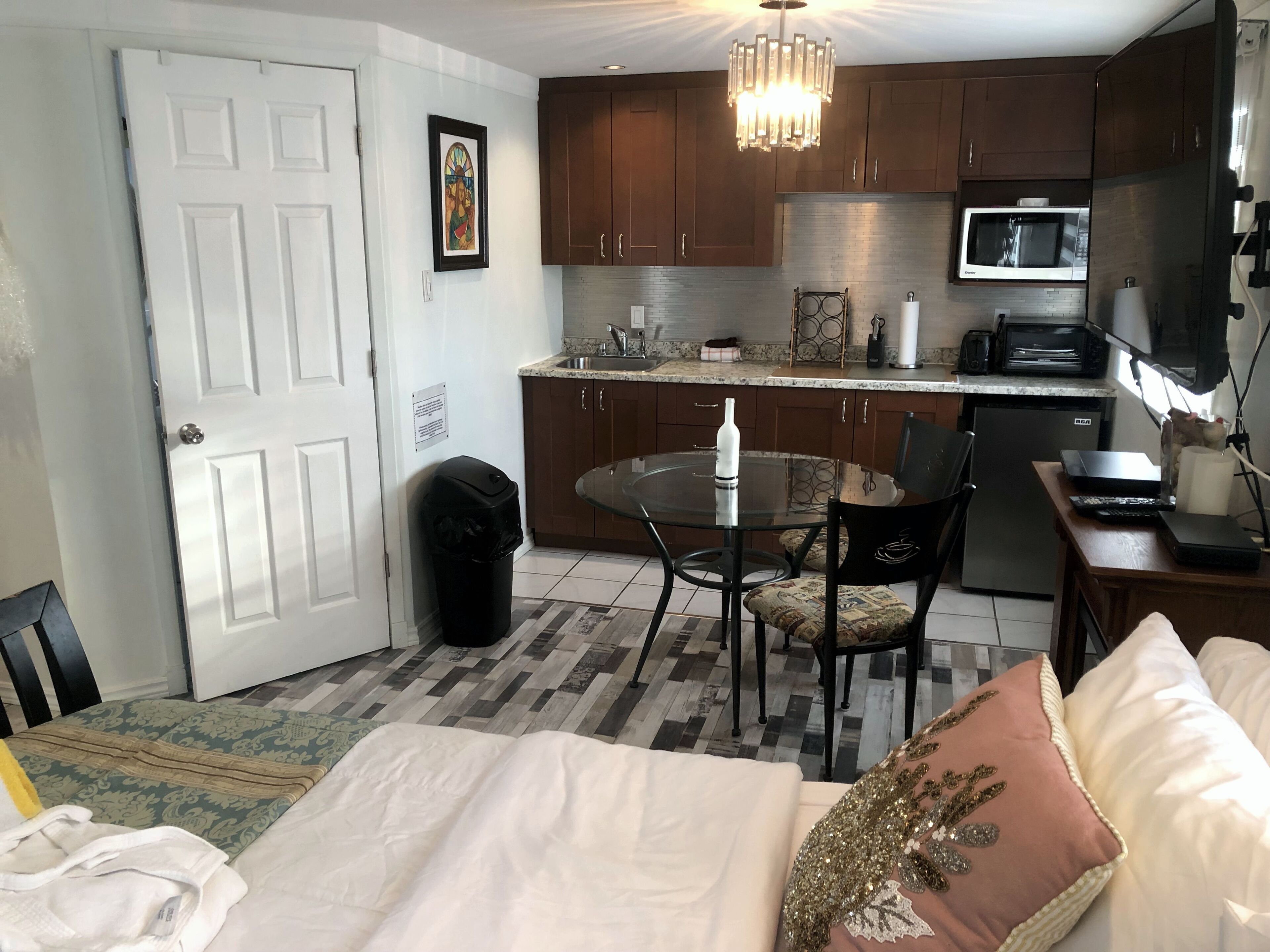 A compact hotel room with a bed, a small dining table, a kitchen area with a microwave, and a trash bin.