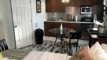 A compact hotel room with a bed, a small dining table, a kitchen area with a microwave, and a trash bin.