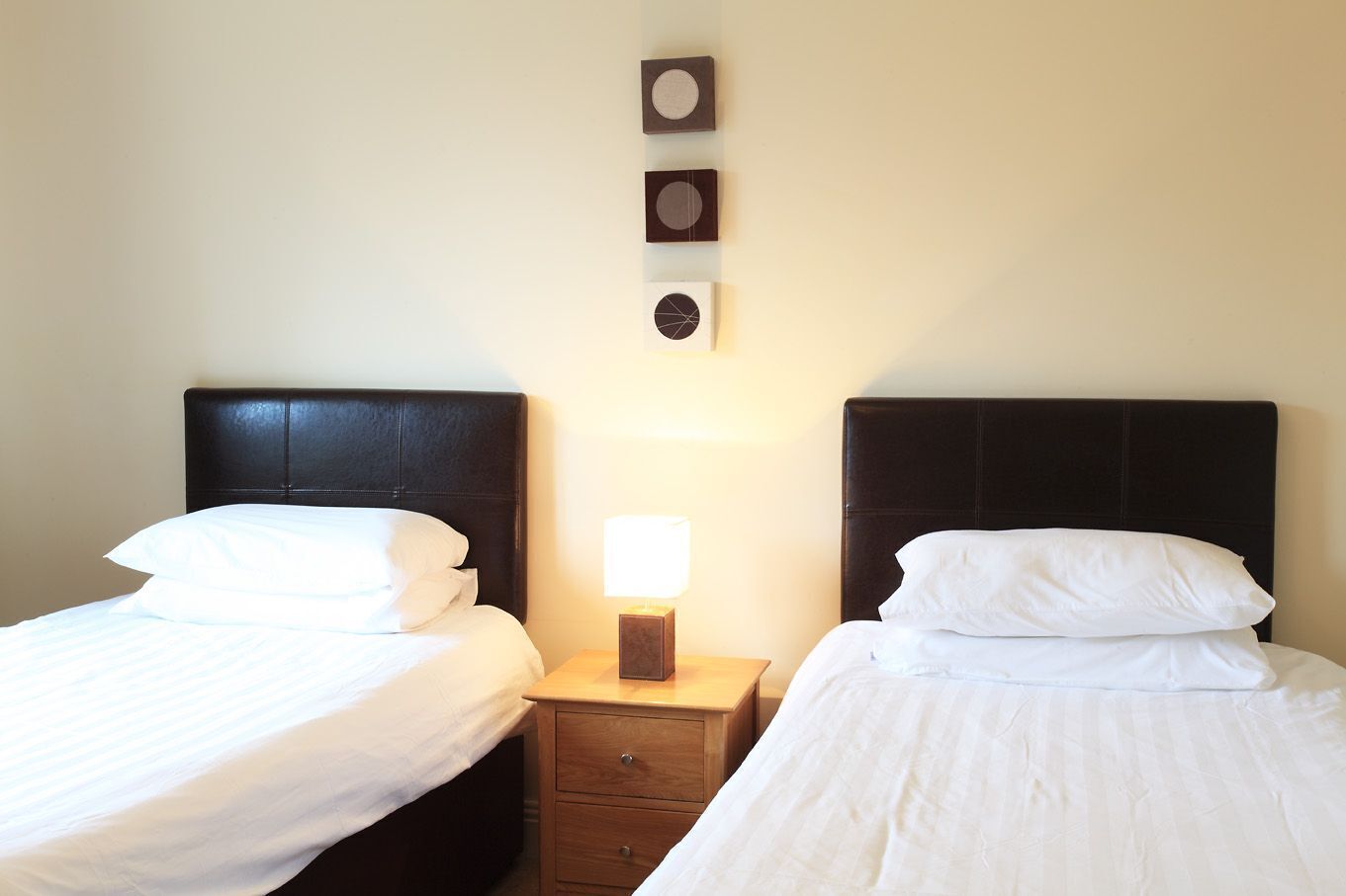 A hotel room with two beds, each with white bedding and a dark headboard. There is a nightstand with a lamp between the beds.