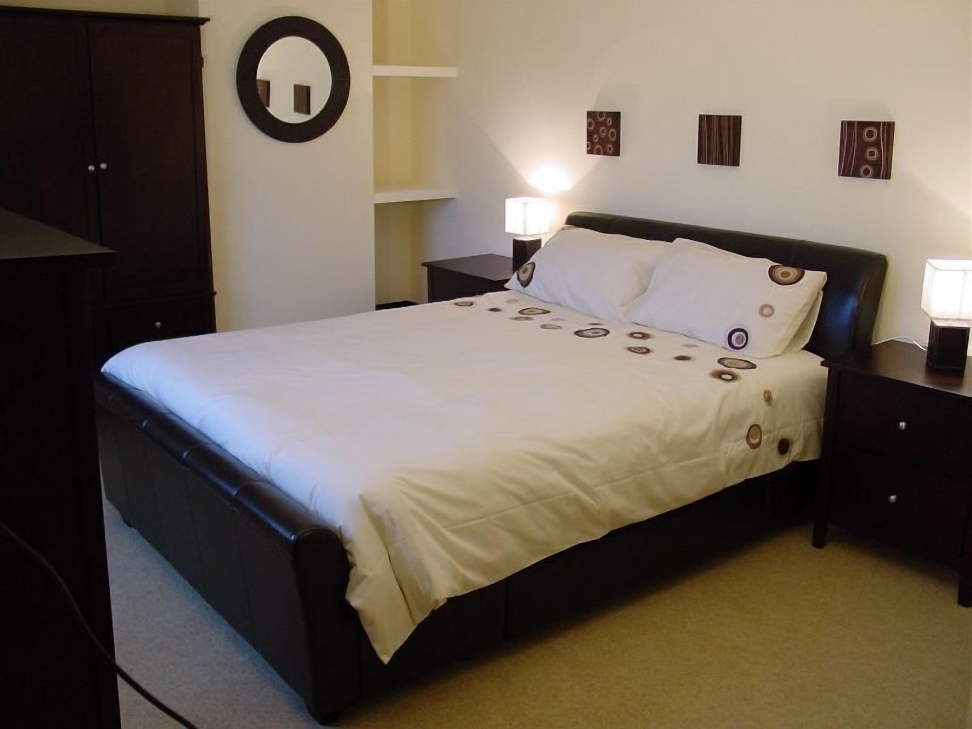 A neatly made bed with a white bedspread and circular pillow designs, flanked by dark wooden nightstands with lamps, in a room with a dark wardrobe and a wall-mounted mirror.