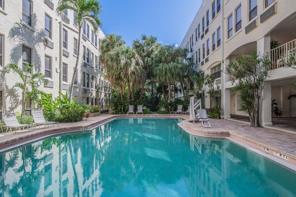 A swimming pool surrounded by palm trees and lounge chairs, with multi-story buildings in the background.