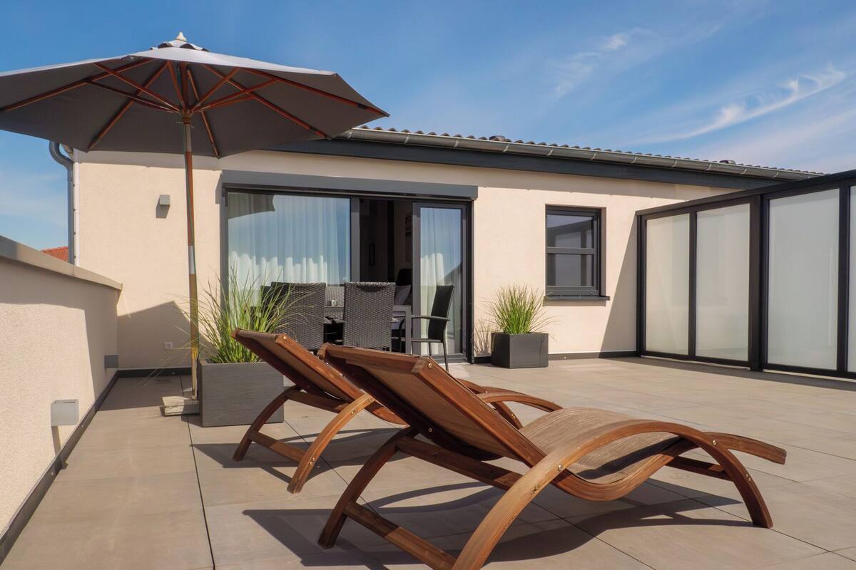 Un espace extérieur moderne aménagé avec deux fauteuils en bois et un grand parasol, situé à côté d’un bâtiment doté de grandes fenêtres et d’une porte-fenêtre coulissante.