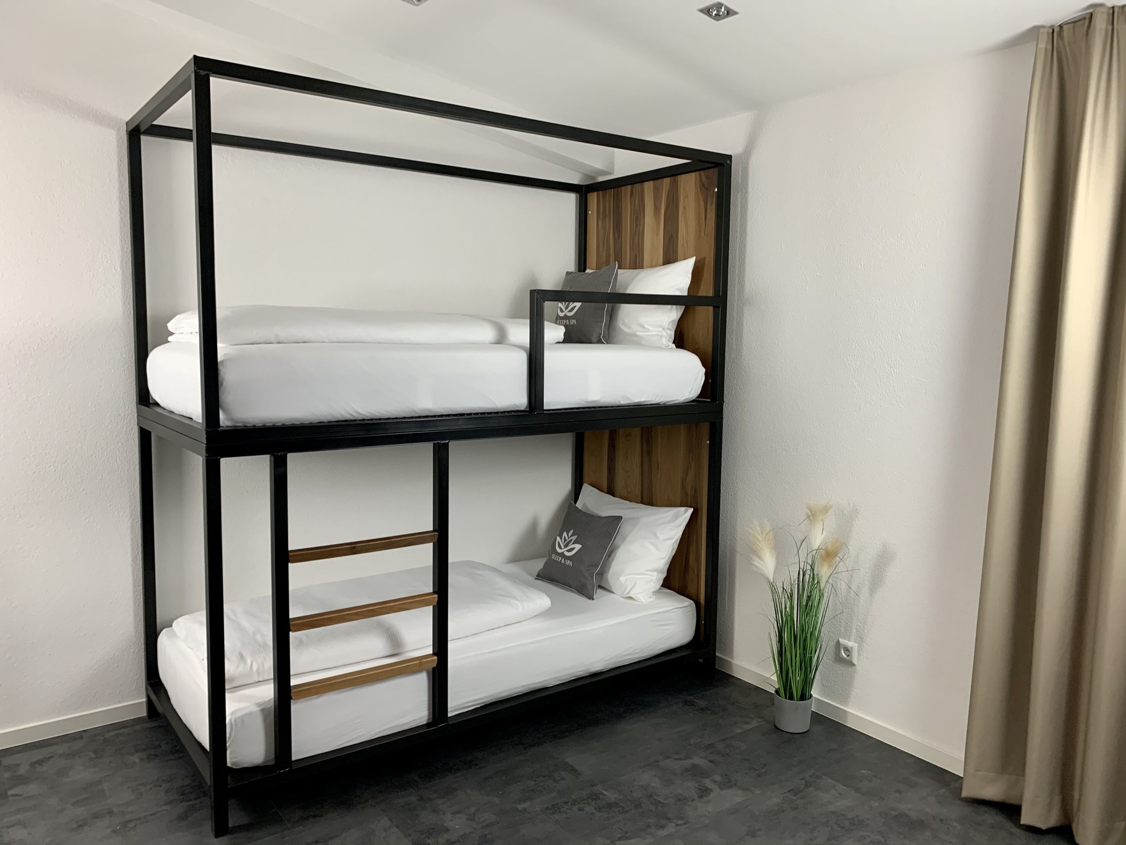 A bunk bed with a wooden frame and white bedding, a small bedside table with a lamp, and a potted plant.