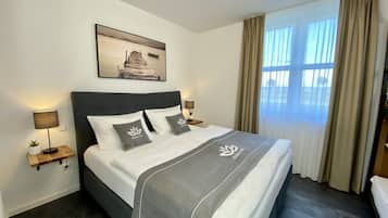 Quarto de hotel com uma cama, duas mesas de cabeceira, um criado-mudo, uma lâmpada, um quadro na parede e uma janela com cortinas.