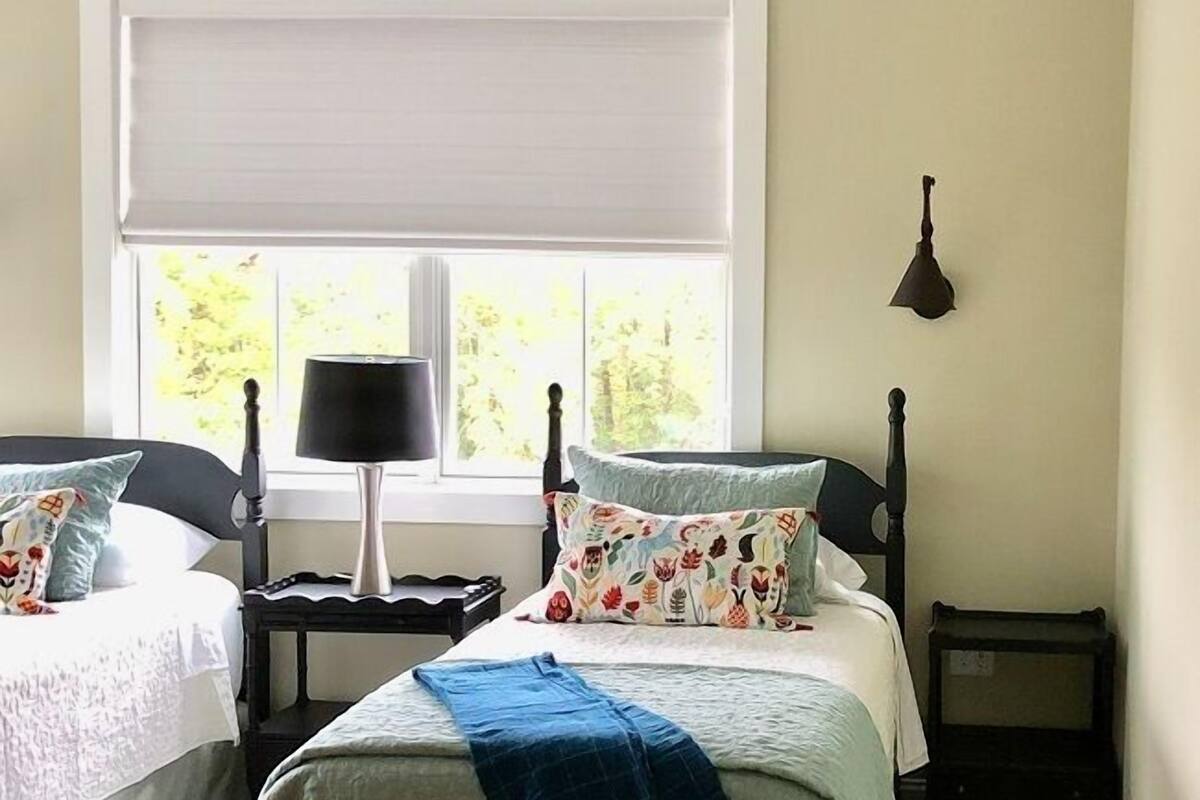 A bedroom with a bed, two bedside tables, a lamp, a window with blinds, and a view of greenery outside.