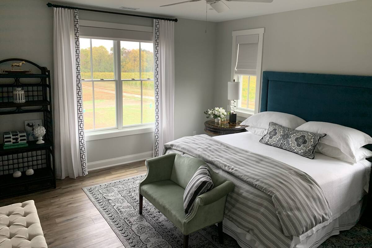 A bedroom with a bed, a chair, a nightstand, a window with curtains, a ceiling fan, and a view of the outdoors.