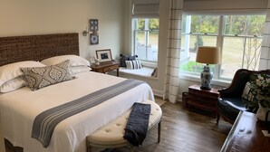 A bedroom with a large bed, a bedside table with a lamp, a window with curtains, and a view of the outdoors. - The Farmhouse (Hammonton)